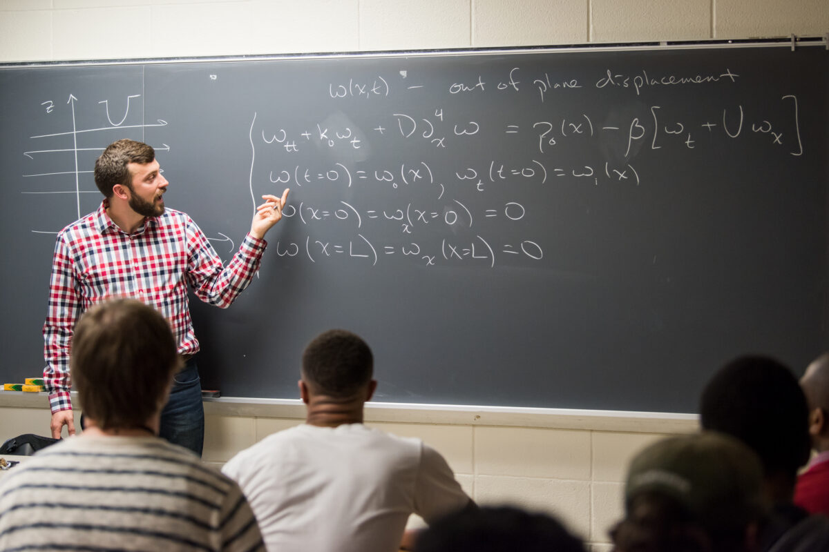 professor points at chalkboard with lots of equations on it; students sit at desks listening