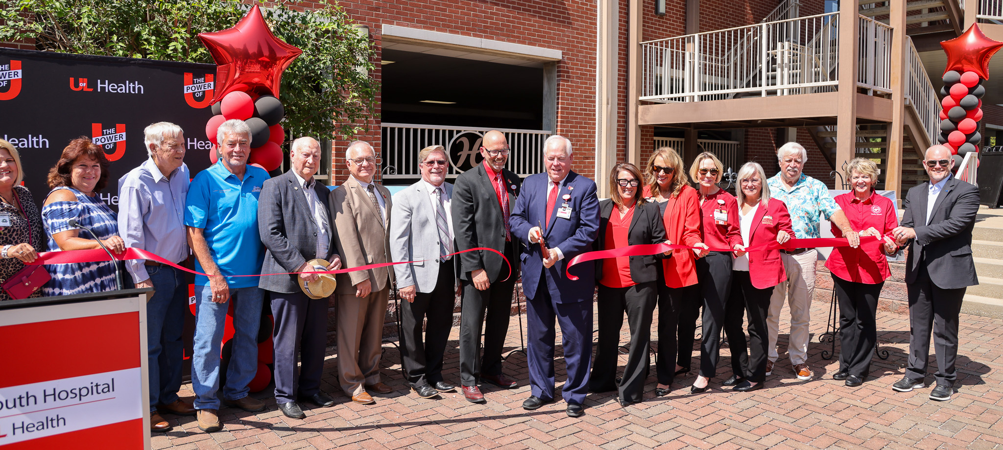 Group of individuals participating in a ribbon-cutting ceremony outside UofL Health – South Hospital Medical Plaza 2, with red and black balloons and banners.