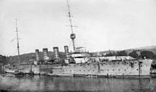 A four-funnelled warship lies at anchor in a harbour.