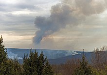 A distant ridge, seen as spring appears to be emerging, with two areas from which white smoke is rising. In the center a large plume of smoke, dark against a cloudy late afternoon sky through which some sunlight is penetrating, rises and bends to the right. In front are some evergreens.