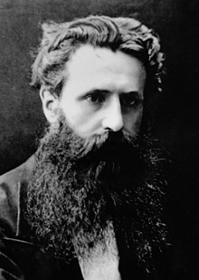 Photographie en noir et blanc d'un homme avec une barbe fournie et une moustache, arborant une coiffure d'époque et un regard sérieux.