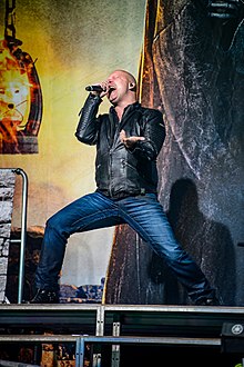 Michael Kiske rozkročmo stojící na vyvýšeném pódiu a zpívající do mikrofonu. Volnou levou rukou gestikuluje.