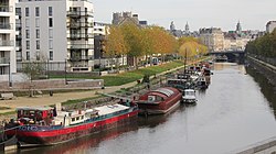 an urban river flowing past modern apartment buildings in the foreground past trees whose leaves are changing with autumn under a bridge and into the city center of Rennes. There are boats docked along the side of the river.