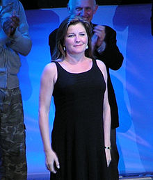 A woman with shoulder length brown hair and a dark dress stands in front of a clapping man while looking forward.