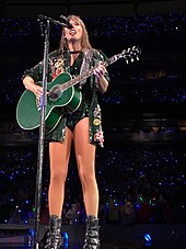 Swift, in a green jacket with lots of detailing and stickers on it, performing a song on a green guitar.