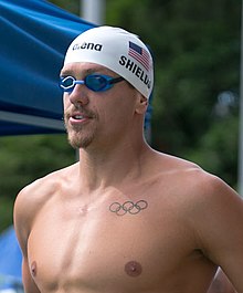 Tom Shields steht unter freiem Himmel neben dem Pool mit Bart, blauer Schwimmbrille und weißer Badekappe mit US-Flagge. Auf seiner Brust hat er die Olympischen Ringe eintätowiert.