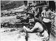 Photograph of a number of uniformed men in a shallow trench, firing rifles and a machine guns towards the left; they appear relaxed and some are smiling.