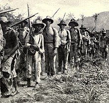 A column of Cuban Liberation Army soldiers marching. Most of the combatants are Afro-Cuban, wear torn clothing, and carry old rifles