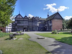 Photo a long, two-storeyed house with exposed wooden beams, many gables and small-paned windows. In the foreground is a lawn and trees are on the left.