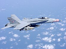 Overall-gray jet fighter, with red, blue and white-tipped nose, is overflying sea and scattered white clouds down below. The aircraft is carrying streamlined external fuel tanks and missiles under its wings, and is heading right.