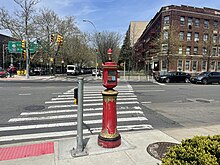 A red New York City Fire Department call box at the intersection of Ocean Parkway and Church Avenue. There is an accessible pedestrian signal to the left and a crosswalk in front of the call box.