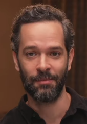 44 year-old man with short black hair and a beard smirking to the left of the camera