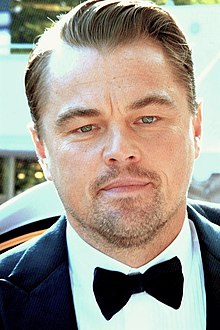 L'acteur Leonardo di Caprio, avec des rides visibles (notamment sur son front), un début de calvitie sur ses tempes, et une barbe taillée. Il est habillé d'un costume avec un nœud papillon.