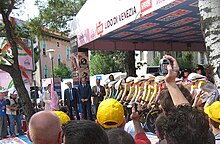 A group of nine cyclists, each wearing identical yellow and white jerseys with black trim, sit crouched over their bicycles in preparation to start a race. Spectators watch on either side of them.