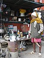 Chaiwala at a stall in Varanasi, a pot of chai is on the stove, and a tray of clean glasses is visible on the table in the background.