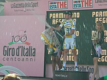 A cyclist in a yellow and white jersey with black trim popping a champagne bottle on a podium, flanked by two beautiful women holding flowers and wine.