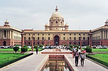 Large building on grassy grounds. A walkway with pedestrians and central reflecting pools leads to the arched entrance. The ground floor is red; the rest of the building is beige. A main cupola is atop the center of the building.