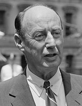 A man with receding dark hair wearing a black jacket, white shirt, and striped tie