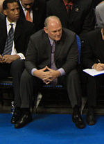 A man, wearing a black suit and a blue tie, is watching the basketball game while sitting courtside.