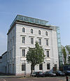 A four-storey building with four bays painted pale grey, with an extra glass storey on the roof and a glass lift on the right side