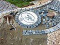 Ein Pflastermosaik in einer Stahlpfanne und die umgebenden Steine werden im Sand verlegt. Das Mosaik symbolisiert die Arbeit des Mitarbeiter des Garten- und Tiefbauamtes und zeigt, wie die Steine mit einem Besetzhammer angedrückt werden.