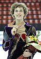 Jeune homme aux cheveux bouclés mi-longs sourit en tenant de sa main droite une médaille et dans sa main gauche un bouquet de fleurs après une compétition sportive