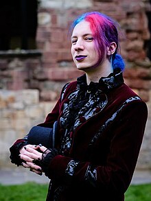 Photograph of me_and, with bright red and blue hair, wearing a black and dark red velvet coat and holding a top hat