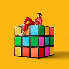 A man is sitting on a rainbow Rubik's Cube against a yellow background.