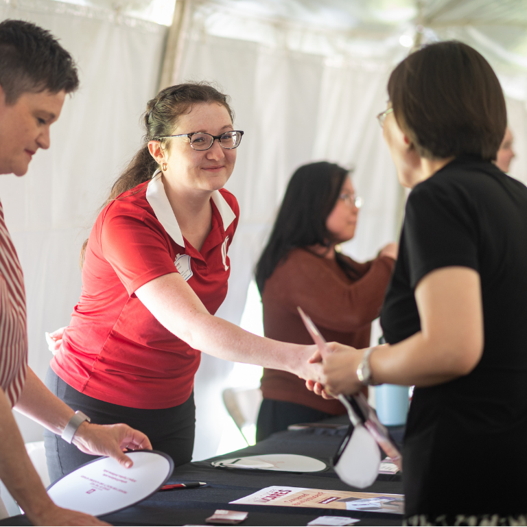 A woman in a red IU branded shirt shakes hands with another woman across a table at the New Faculty Orientation Picnic and Resource Showcase. 