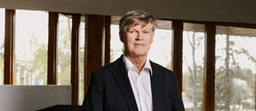 Tapani Vuorinen in a black suit, facing camera in front of large windows.