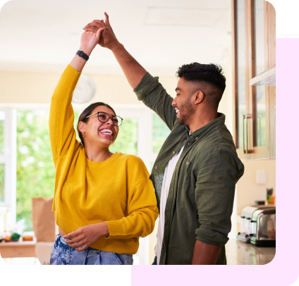 A couple, hand in hand, laugh as they dance together in their kitchen.