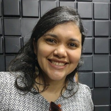 Angela é uma jovem, com cabelo liso na altura dos ombros, preto e com uma mecha grisalha. Ela olhos pretos e está sorrindo e olhando para a foto. Ela está vestindo uma camiseta branca com detalhes geométricos pretos. Ela tem um óculos de sol pendurado na gola da camiseta.