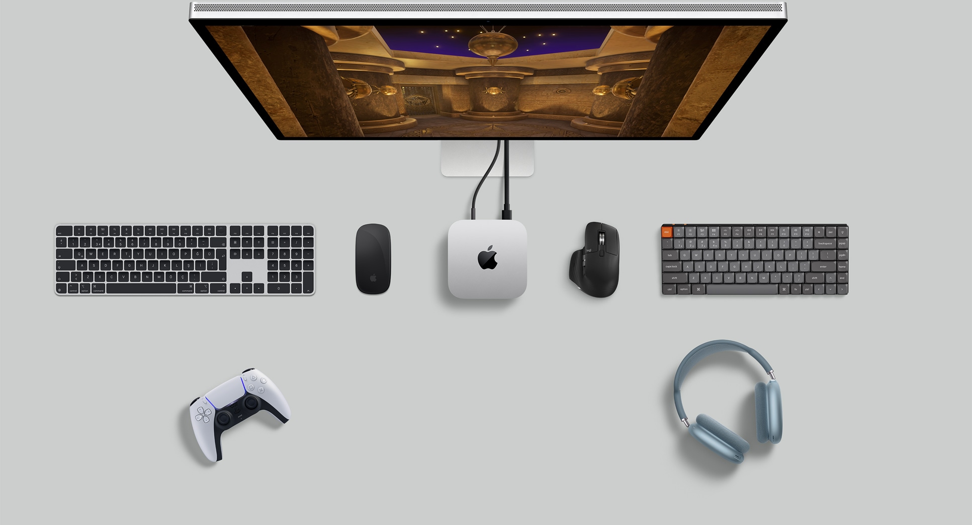 Gümüş rengi Mac mini, bir ekran, kablosuz klavyeler, kablosuz mouse’lar, bir oyun kumandası ve AirPods Max’in bulunduğu masa düzeninin üstten görünümü