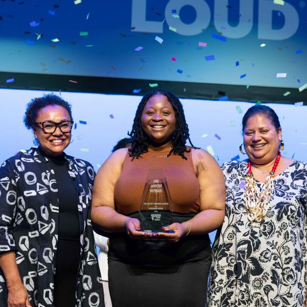 Three women stand smiling on a stage as confetti fall around them, the young woman in center holds a trophy 