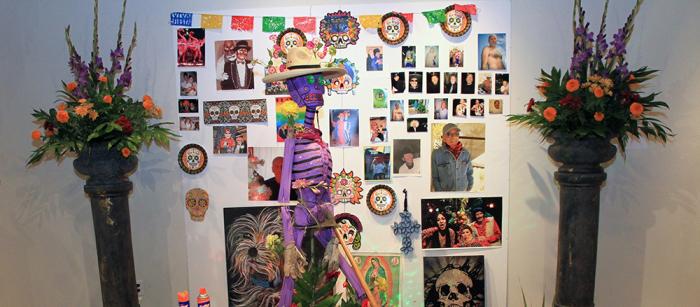 An altar for a Day of the Dead celebration with a purple skeleton, orange flowers, and a poster with photos on it. 