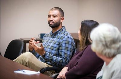 A young black man with a shaved head and beard wearing a button-down shirt and pants talks to a group of people.