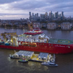 A large red ship is floating in a river with a city skyline behind it.