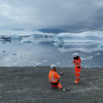 Two people standing in front of an icy lake with snow covered mountains behind