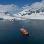 A red ship sailing through a flat ocean, with snow covered mountains behind