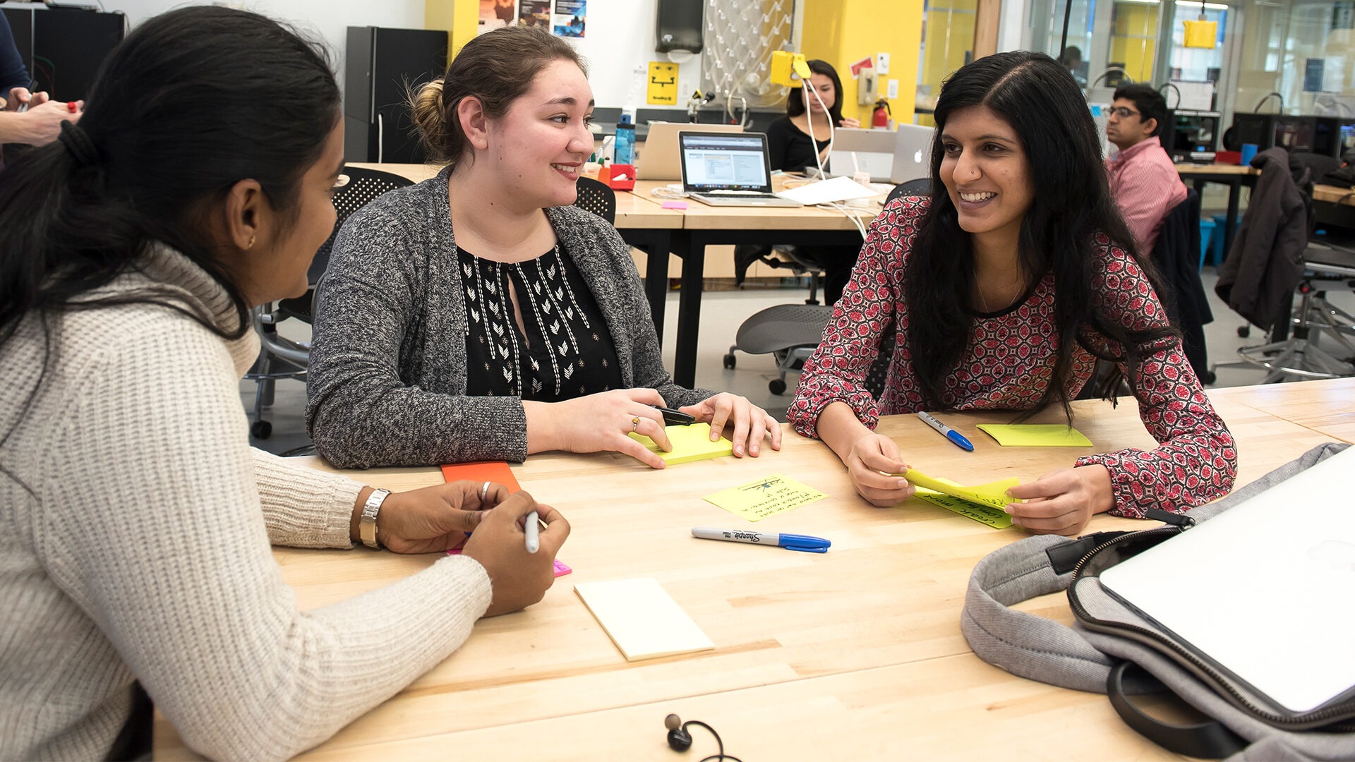 Three female students sit at a table in the design studio and discuss something during class.