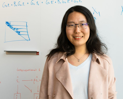 Headshot of Yue Qin standing in front of a whiteboard with equations written on it.