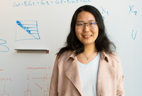 Headshot of Yue Qin standing in front of a whiteboard with equations written on it.