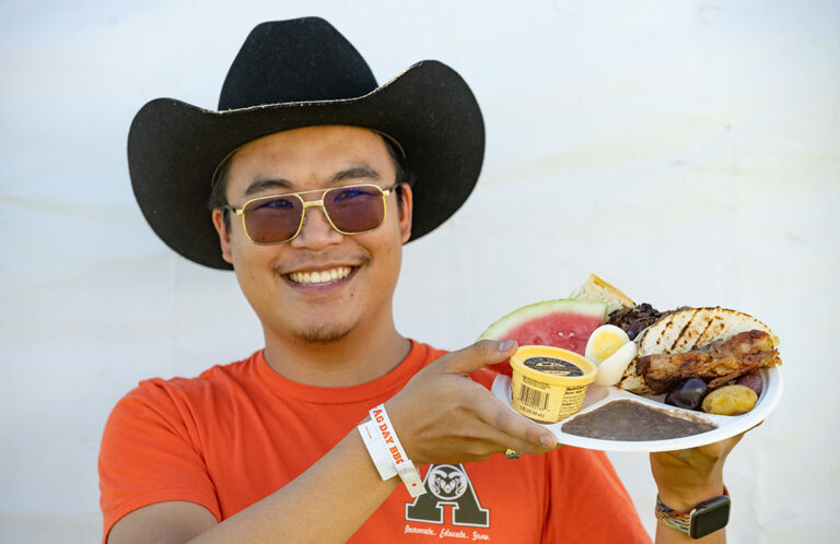 Photo of person in cowboy hat, orange shirt with "A" logo holding a plate of BBQ food.