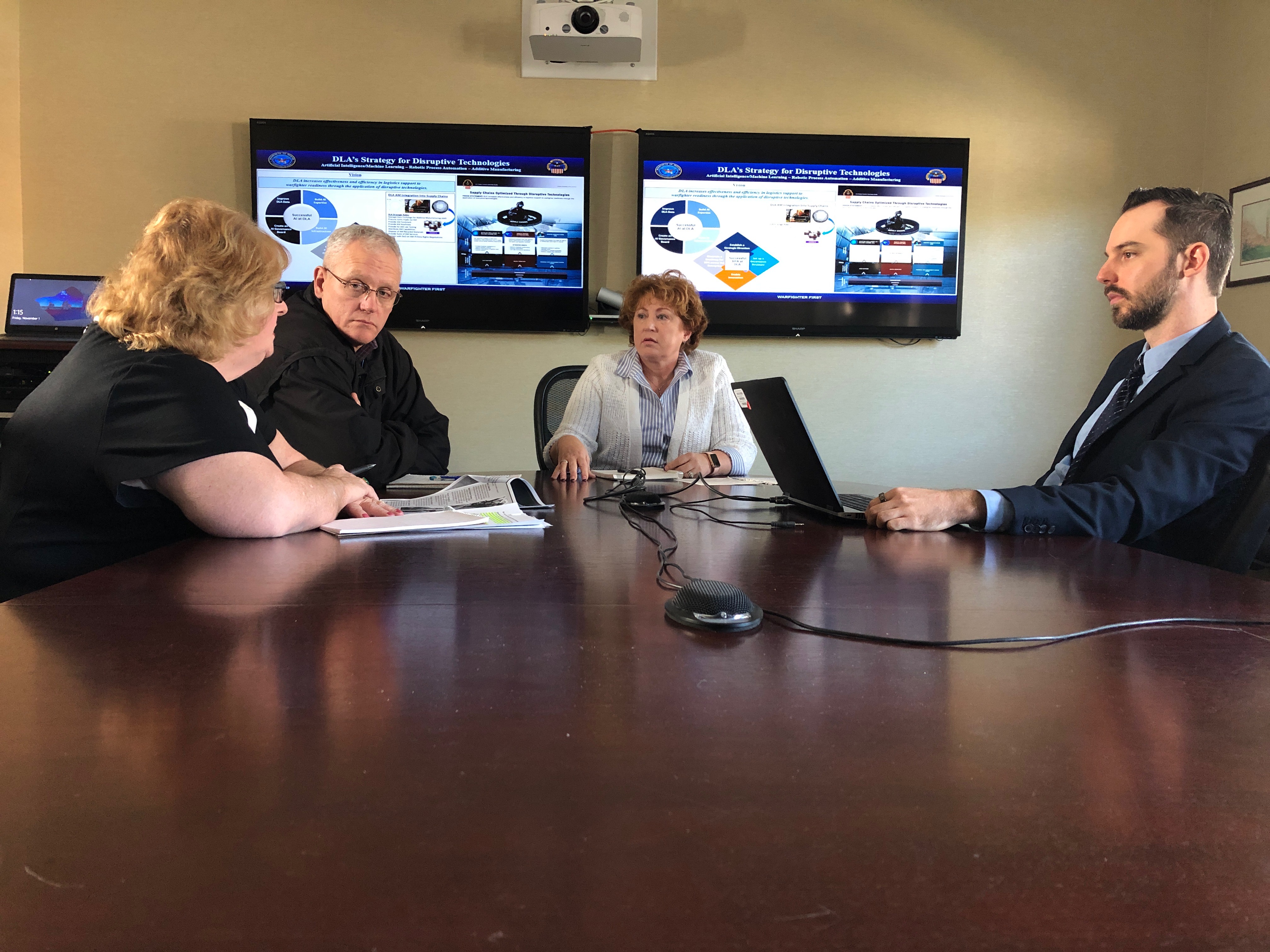 Coworkers have a discussion around a conference table with charts displayed on screens behind them