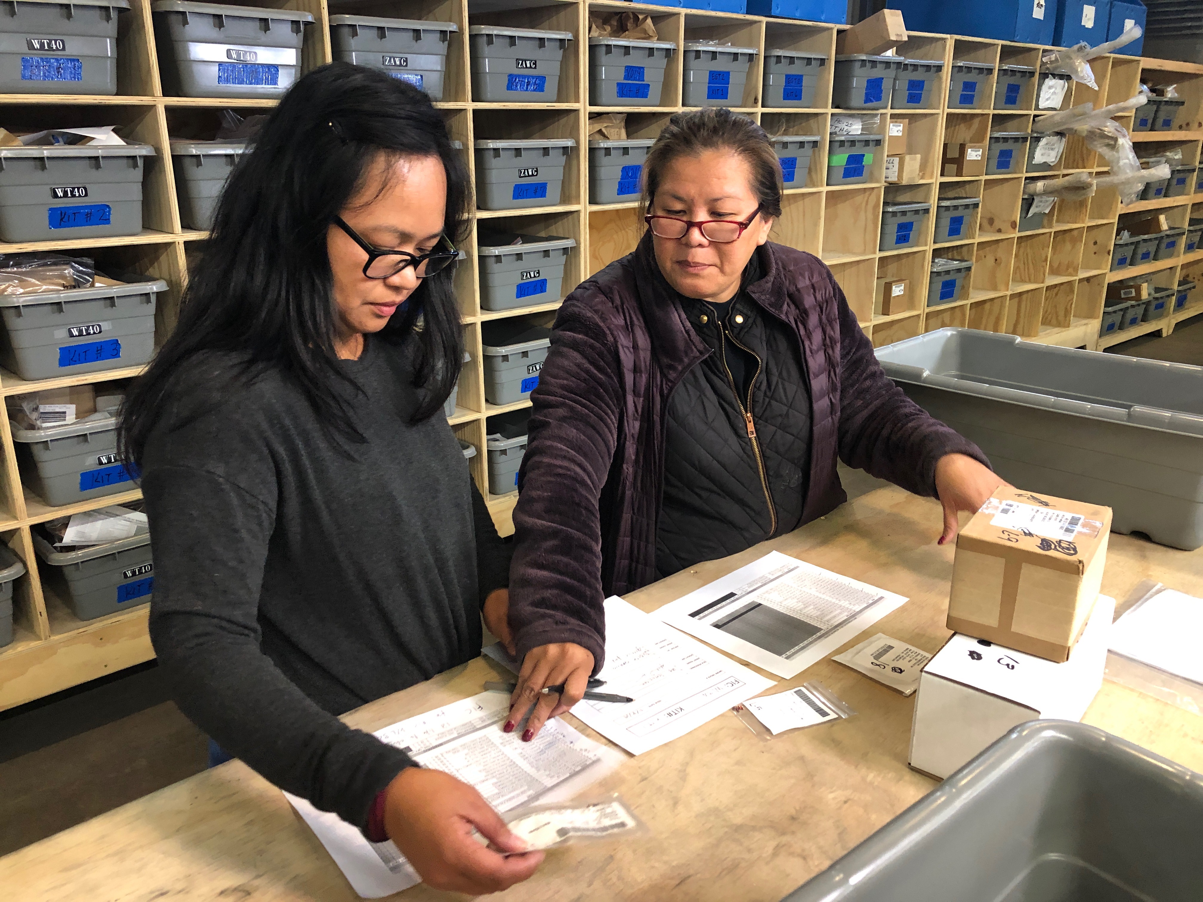 Two workers in a warehouse examine paperwork with shelves of storage bins in the background