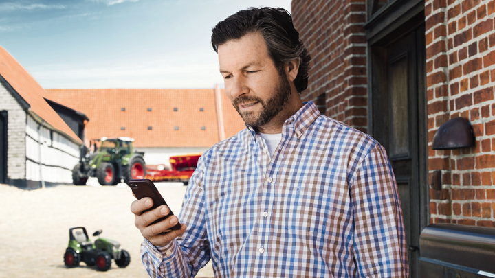 A farmer stands in front of his farm holding a mobile phone.