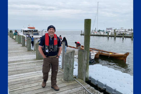 A man wearing a beanie and life jacket stands on a dock. Behind him are several boats, including a canoe.