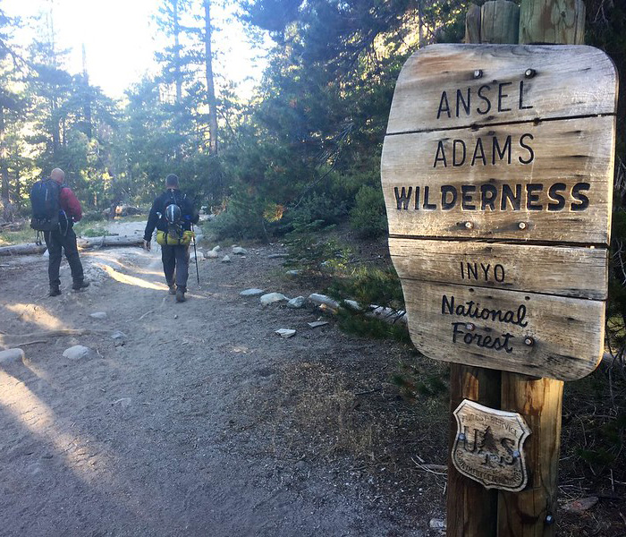 Two hikers walking down a wilderness trail just past a sign that reads, Ansel Adams Wilderness - Inyo National Forest.