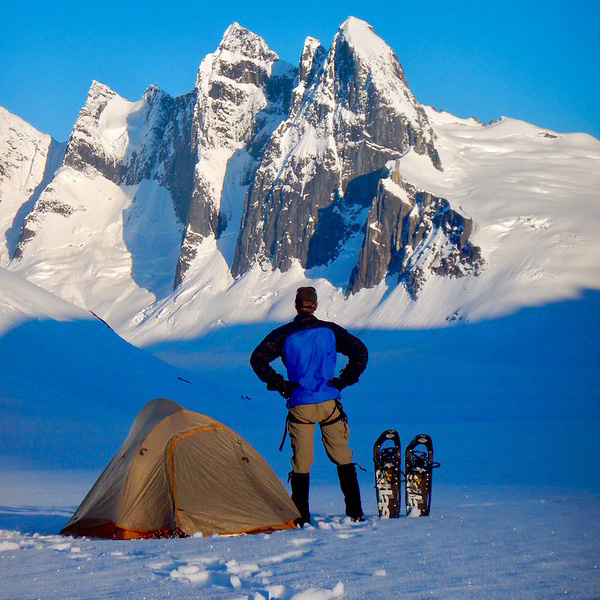 A person standing in the snow, next to a tent and a pair of snow shoes, facing away from the camera, looking up at the rocky, snow covered peaks of the Mendenhall Towers.