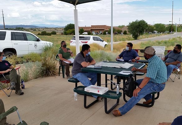 A group of people wearing masks sit at a table under a picnic pavilion.